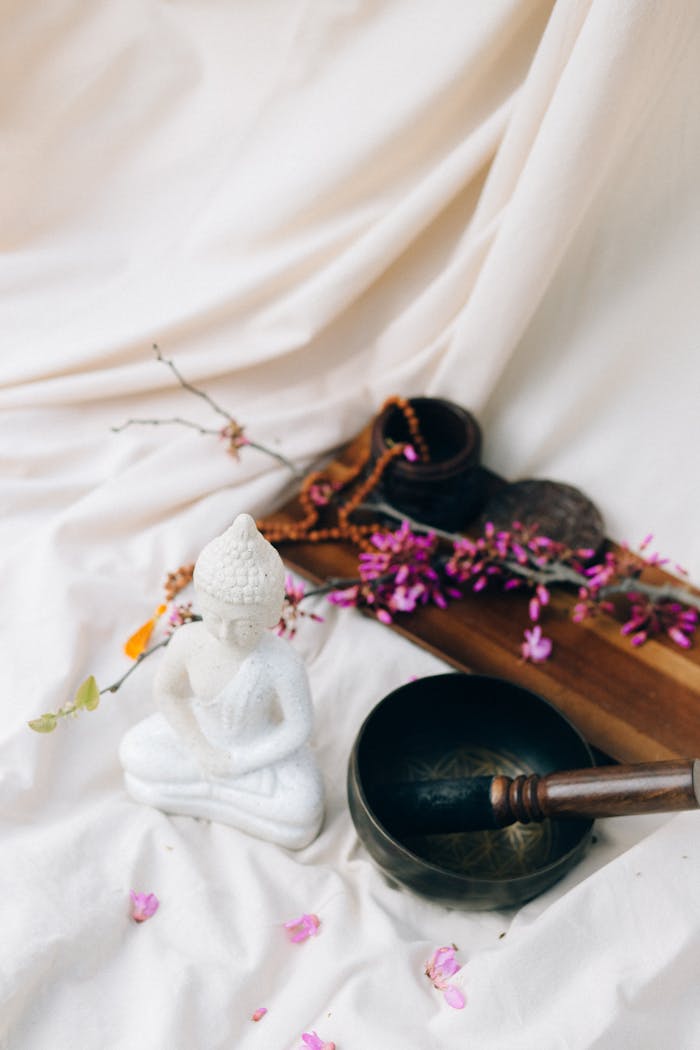 A serene meditation setting featuring a Buddha statue, singing bowl, and pink flowers on a draped fabric surface.